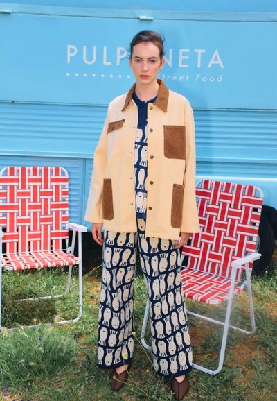 Beige knäppt jacka med bruna mockapatchar, tillsammans med lösa blå byxor med vita fiskmönster; modellen står på grönt gräs.