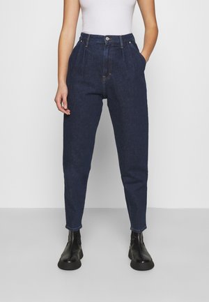 Femme portant un jean taille haute bleu foncé fuselé, un haut blanc sans manches et des bottines noires, debout les mains dans les poches.