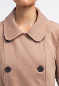 Nærbillede af en person iført en beige dobbeltradet frakke med bred krave og sorte knapper, der viser hals og øverste del af brystet.