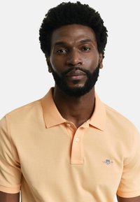 Lys oransje polo skjorte laget av teksturert stoff, med krage og tre knapper ved halsen. Lite logo-patch på venstre bryst.