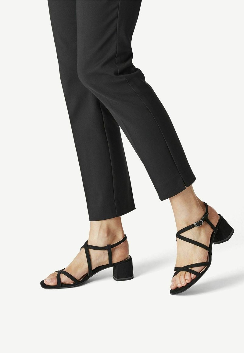 Pieds portant des sandales noires à brides avec des talons blocs bas, assorties à un pantalon noir raccourci sur un fond clair.