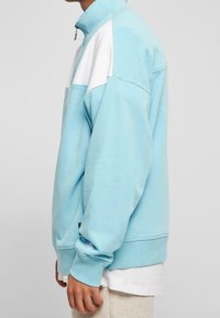 Ljusblå sweatshirt med hög krage, vita detaljer, avslappnad passform, ribbade ärmmuddar och en liten logotyp på sidan. Mjukt, strukturerat material.