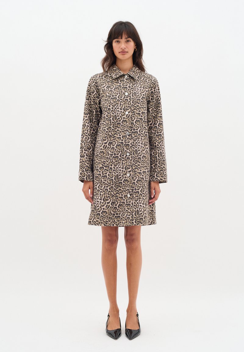 Leopardmönstrad kappa i textil, med knappknäppning fram, krage och långa ärmar, skuren ovanför knäet. Svarta klackskor bärs.