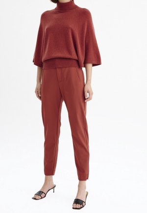 Woman wearing rust-colored loose turtleneck sweater, matching tapered pants, and black open-toe heeled sandals standing against white background.