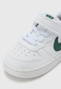 Λευκό αθλητικό παπούτσι με δερμάτινο πάνω μέρος, πράσινο σήμα Nike, διάτρητο μπροστινό μέρος και συνδυασμό κορδονιών και ιμάντα με αυτοκό�λλητο για κλείσιμο.