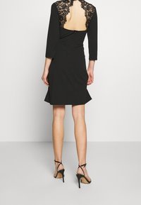 Czarna sukienka z koronkowym tyłem, rękawami do trzech czwartych, dopasowaną sylwetką i rozkloszowanym dołem. Noszona z czarnymi sandałami na obcasie.