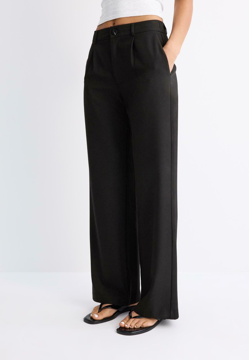 Femme portant un pantalon noir taille haute à jambes larges avec plis devant, poches latérales et sandales plates noires à lanière entre les orteils sur fond blanc.