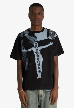 Giovane uomo che indossa una t-shirt nera oversize con figura grafica grigio chiaro e la scritta "acupuncture", abbinata a jeans in denim chiaro.