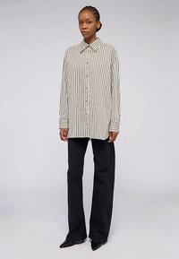 Gestreiftes Hemd in Beige und Schwarz mit Kragen und langen Ärmeln, kombiniert mit locker sitzenden schwarzen Hosen und spitzen Schuhen.