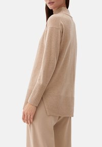 Femme portant un pull beige en maille avec poignets et ourlet côtelés, associée à un pantalon beige, debout devant un fond clair uni.