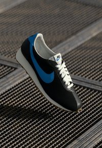 Melna un zila Nike tenisa kurpe ar baltajiem sējumiem, kurai ir ribota tekstūra un izteikts zils svižs, uz metāliski spīdīga fona.