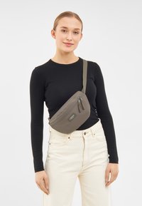 Szürke, vízálló válltáska cipzáras zárással és állítható pánttal, fekete felsőn és fehér farmernadrágon viselve.