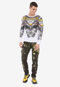 Langärmliges weißes Hemd mit Adlergrafik und gelben Akzenten, kombiniert mit schwarzen Hosen im Batikmuster und metallischen Nieten-Details. Gelbe Sneakers.