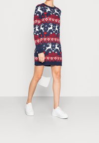 Femme portant une robe-pull de Noël bleu marine avec des rennes blancs, des cœurs rouges, des sapins et le texte "Joyeux Noël", associée à des baskets blanches.