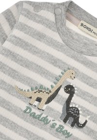 Grau gestreifter Overall mit gestickten Dinosauriern in Beige und Schwarz. Druckknöpfe am Halsausschnitt, mit dem Text "Daddy's Boy" in Grün.