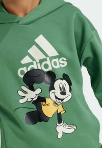 Zelená mikina s kapucí s velkým černo-žlutým motivem Mickey Mouse a bílým logem Adidas. Měkká látka, volný střih a kapuce se šňůrkou.
