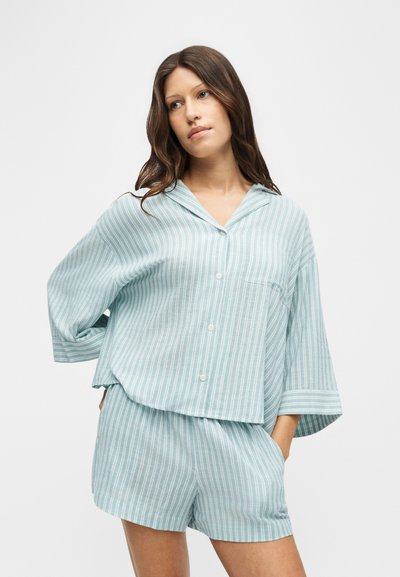 Jeune femme portant une chemise ample rayée bleu clair et blanc à boutons et un short assorti, debout avec une main dans la poche.