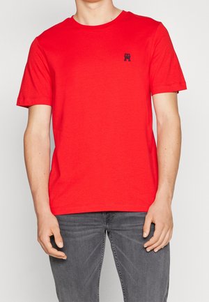 Mann trägt ein rotes T-Shirt mit kurzen Ärmeln und kleinem dunkelblauem Logo auf der Brust, kombiniert mit grauen Jeans, steht vor einem einfarbigen Hintergrund.