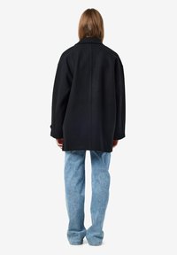 Schwarzer übergroßer Wollmantel mit Reverskragen, entspannter Schulterpartie und Rückennahtdetails, kombiniert mit hellblauen weit geschnittenen Jeans.