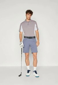 Golfový outfit so šedou a bielou polokošeľou s krátkymi rukávmi, doplnený svetlomodrými šortkami. Doplnky zahŕňajú čierny opasok a bielu golfovú rukavicu.
