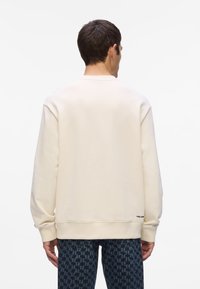 Sudadera crema con cuello redondo y puños de canalé. Textura suave, corte holgado, con un pequeño logo de la marca en el dobladillo.
