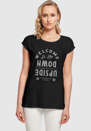 Musta puuvillapaita, jossa on harmaata printtiä, jossa lukee "TERVETULOA ALAS PUOLELLE", rento ja väljä istuvuus sekä käännetyt hihat.