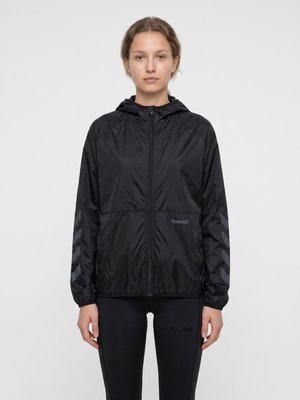 Jeune femme portant une veste noire à capuche zippée avec des motifs chevrons sur les manches et des leggings noirs, debout devant un fond uni.