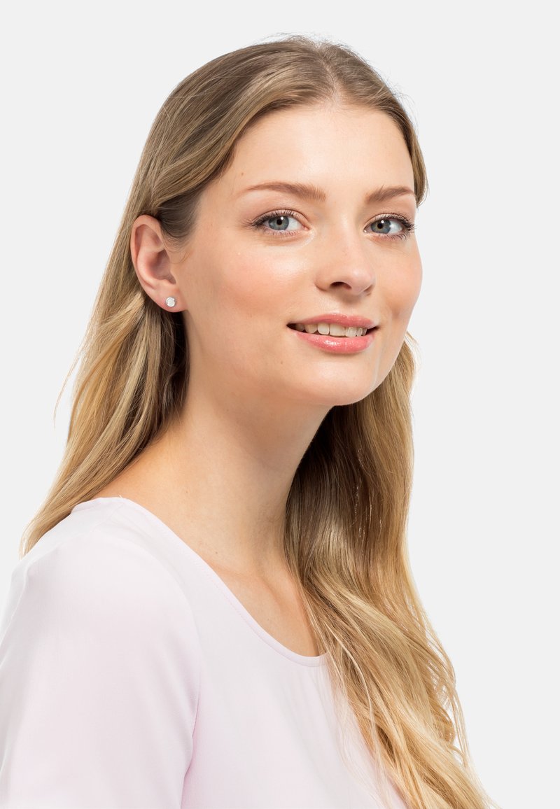 Weiße Steckerohrringe, getragen von einer Frau mit langen, glatten, blonden Haaren. Die Ohrringe sind rund und schlicht und passen gut zu einem hellrosa Oberteil.