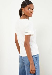 Donna con capelli scuri raccolti in una coda di cavallo bassa, che indossa orecchini a forma di conchiglia dorati, una maglietta bianca a maniche corte e jeans blu, che guarda oltre la spalla.