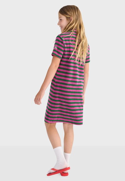 Fille aux longs cheveux blonds portant une robe rayée rose et verte, des chaussettes blanches et des tongs rouges, debout et souriant de profil.