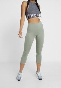 Šedý sportovní croptop s páskem Nike Pro, v kombinaci se světle zelenými capri legínami s vysokým pasem. Modelka má na sobě šedé a modré běžecké boty.