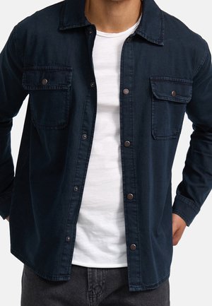 Mand iført en åben mørkeblå langærmet skjorte over en hvid t-shirt og sorte bukser, hænderne hvilende på hofterne.