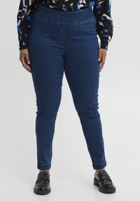 Pantalones de mezclilla azul oscuro con una cintura alta, que presentan un sutil detalle en las costuras y un corte ajustado, combinados con zapatos negros de tipo slip-on.