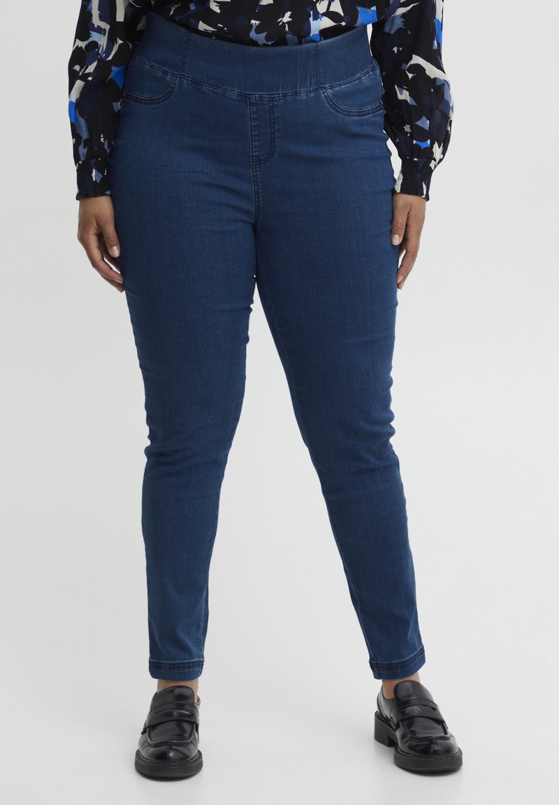 Pantalones de mezclilla azul oscuro con una cintura alta, que presentan un sutil detalle en las costuras y un corte ajustado, combinados con zapatos negros de tipo slip-on.