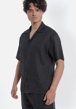 Uomo con capelli ricci che indossa una camicia nera a maniche corte con bottoni e pantaloni neri, in piedi davanti a uno sfondo bianco uniforme.