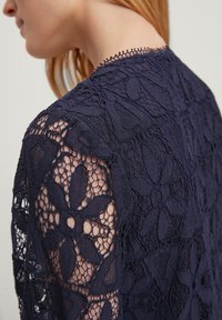 Top en dentelle bleu marine avec des motifs floraux, mettant en avant des détails fins, une texture tissée et une bordure festonnée le long de l'encolure et des manches.