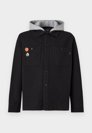 Jeune homme portant une tenue superposée avec une veste noire, un sweat-shirt gris avec un logo triangulaire, une chemise à carreaux et un collier de perles, debout devant un fond blanc.