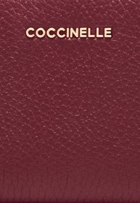 Superficie in pelle testurizzata borgogna con lettere metalliche dorate che spellingano "COCCINELLE" centrate vicino alla parte superiore.