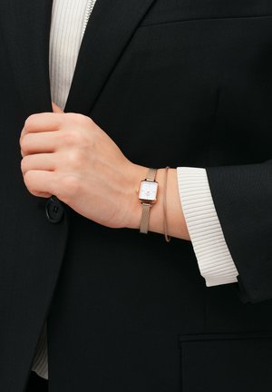 Rose gold mesh watch with a rectangular white face. Slim wrist, wearing a brown elastic bracelet, against a dark blazer. White ribbed sleeve visible.