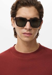 Tortoiseshell sunglasses with dark lenses, rectangular frames, and subtle pattern detail. Worn by a person in a red crew-neck t-shirt.