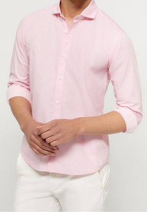 Man draagt een lichtroze overhemd met lange mouwen, waarvan de mouwen zijn opgerold, en een witte broek, handen gevouwen voor zich.