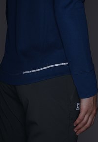 Marinblå långärmad topp med texturerad tyg och reflekterande detaljer i fållen. Kombinerad med grå byxor med en logotyp.