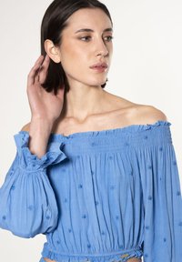 Lichtblauwe off-the-shoulder blouse met smockdetails, geruite manchetten en geborduurde bloemenpatronen. Zacht, vloeiend materiaal.