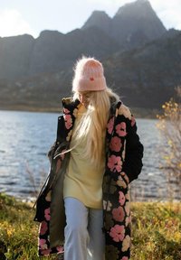 Dlhý čierny kabát s kvetinovým vzorom, svetlý sveter, modré džínsy a ružová pletená čiapka. Vlasy sú dlhé a voľné, stojí pri jazere s horami.