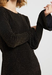 Femme portant un haut en tricot côtelé noir avec des paillettes dorées et de longues manches cloche, levant un bras devant un fond uni.