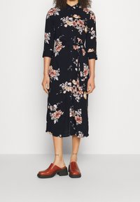 Květinové midi šaty v černé barvě s růžovými a žlutými květinami, zapínání na knoflíky v přední části, límec a pásek v pase. Spárované s hnědými dřeváky.