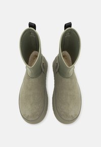 Paire de bottines en daim vert olive avec doublure en polaire, fermeture éclair sur les côtés et languettes noires à l'arrière, vues de dessus.