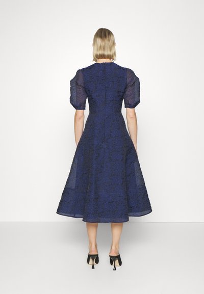 Vestido de renda azul-marinho com mangas curtas e bufantes, corpete justo e saia rodada, apresentando um padrão floral texturizado e comprimento midi.