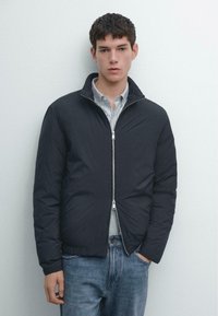 Młody mężczyzna z krótkimi ciemnymi włosami, ubrany w czarną kurtkę na zamek błyskawiczny, nałożoną na pasiastą koszulę i niebieskie dżinsy, stojący na tle jednolitej białej ściany.