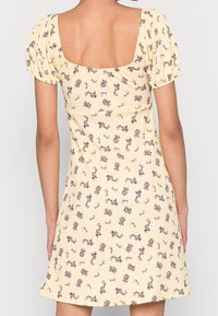 Robe à fleurs jaunes avec manches courtes bouffantes, décolleté carré et un tissu texturé présentant de petites fleurs violettes et des feuilles vertes.